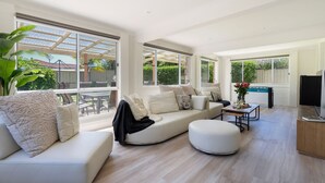 A spacious living room with a large sofa, a round ottoman, and a coffee table. There are multiple windows providing natural light and views of the outdoors. - Marilyn's - 180 degree views of Jervis Bay (Callala Beach)