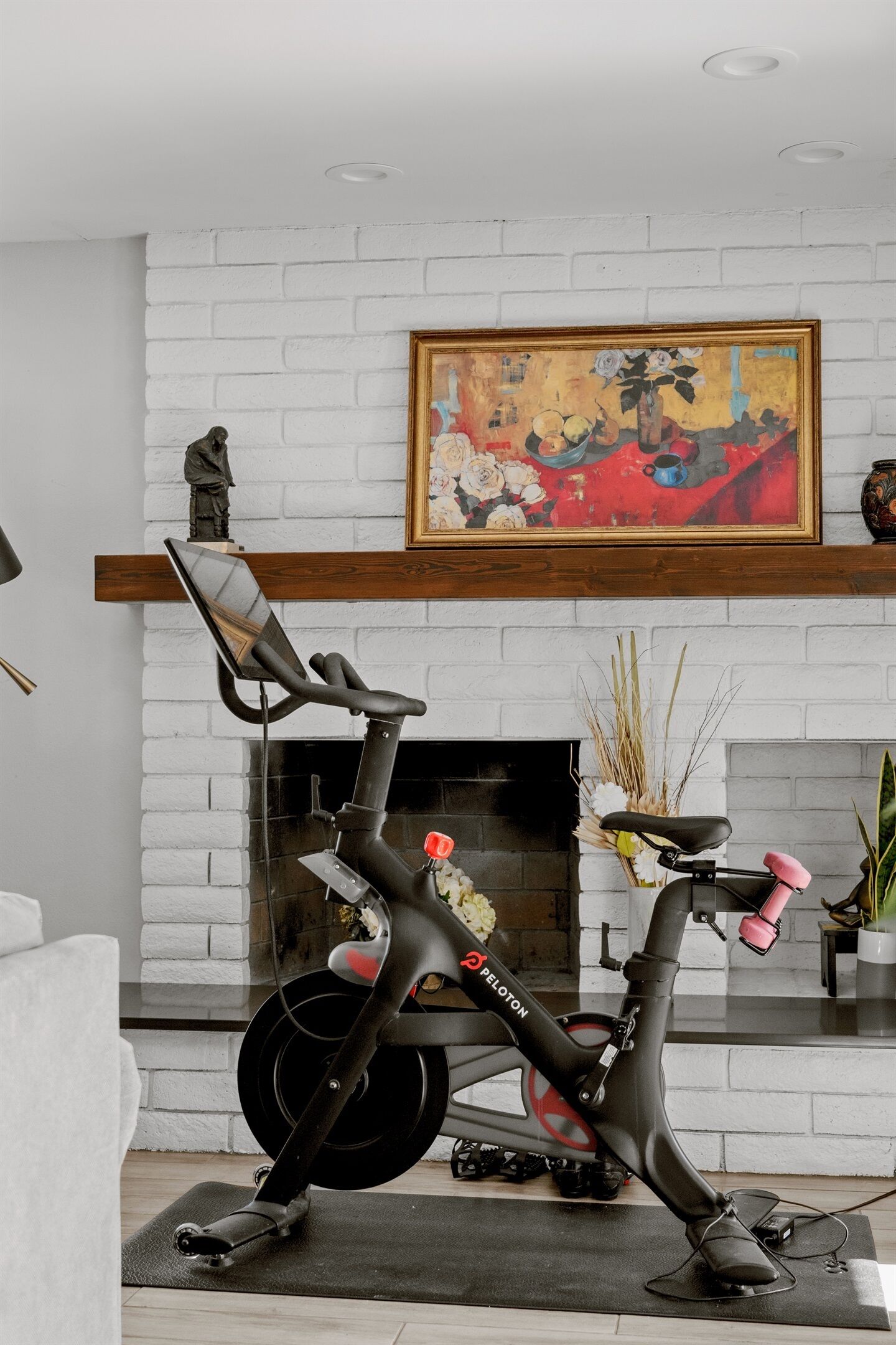 A Peloton exercise bike in a living room with a white brick fireplace, a painting above the mantel, and a small table with a vase of dried plants.