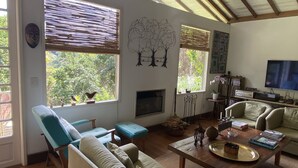 A spacious living room with a high ceiling, wooden beams, a fireplace, and large windows with blinds. - Summer house, surrounded by trees, rural setting with city amenities. (Petrópolis)