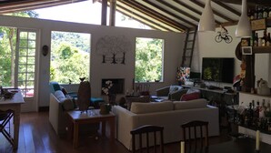 A spacious living area with a large sofa, a dining table, and a kitchen area with a bar. There are large windows providing a view of the outdoors. - Summer house, surrounded by trees, rural setting with city amenities. (Petrópolis)