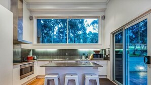 A modern kitchen with a large island, white bar stools, and a view of trees through the window. - Golfview Lorne - For all seasons (Lorne)