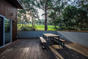 Una terraza de madera con una mesa y bancos, con vista a una zona verde con árboles.