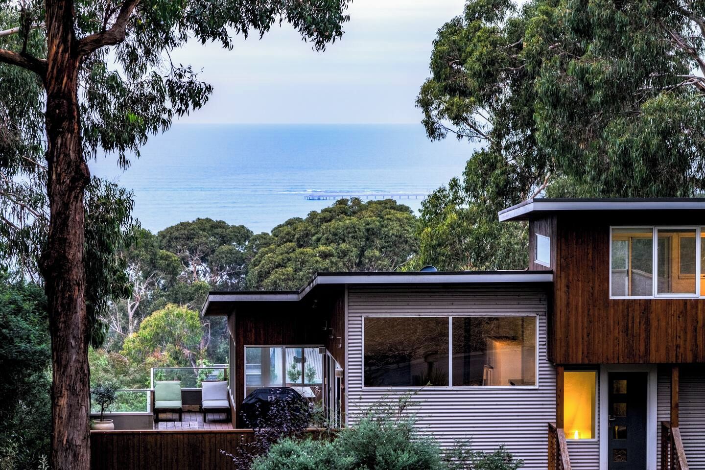 A modern two-story house with a deck, large windows, and a view of the ocean.