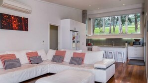 A modern living room with a sectional sofa, a kitchen area, and a large window offering a view of trees. - Golfview Lorne - For all seasons (Lorne)