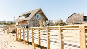 Et træhus ved stranden med skrå tag, en veranda og en trappe til indgangen.