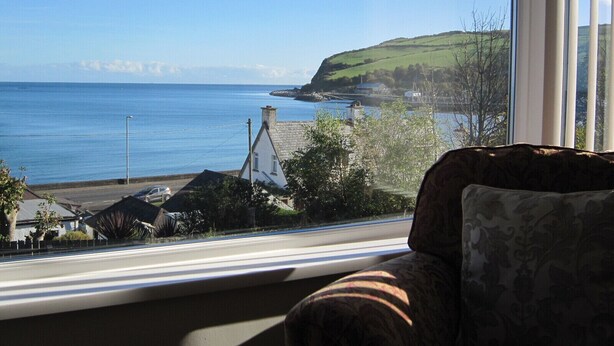 Une vue depuis une fenêtre donnant sur un paysage côtier, avec une chaise et un coussin au premier plan.