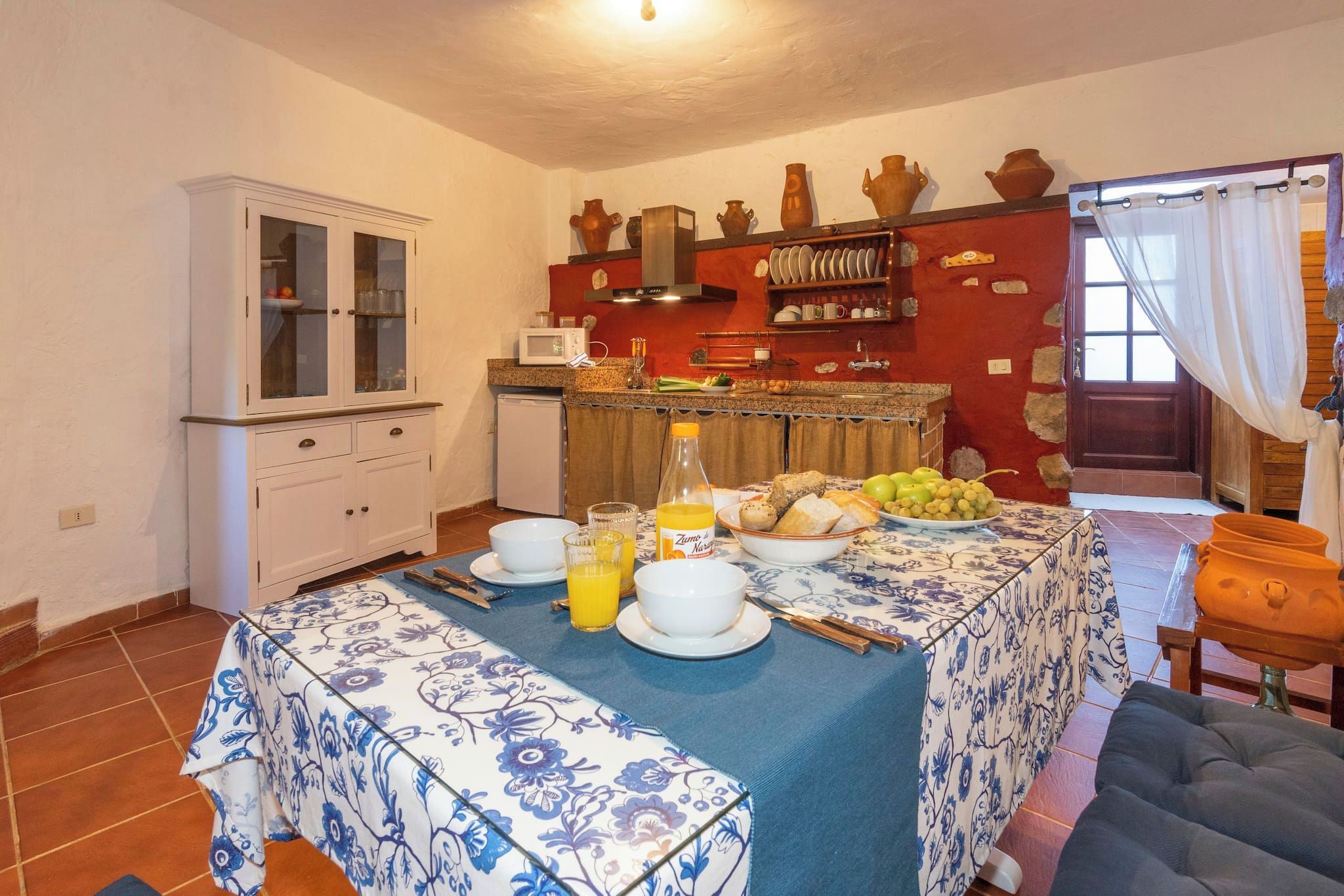 A dining area with a table set for breakfast, a kitchen in the background, and a red wall with decorative pottery.