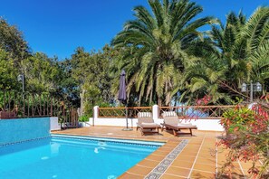 Ein Swimmingpool mit Liegestühlen und einer gefliesten Terrasse, umgeben von Palmen und blauem Himmel.