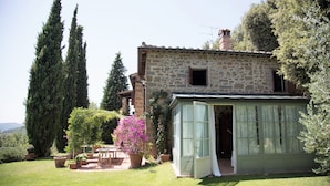 Une maison en pierre dotée d’une véranda, entourée de plantes en pot et de grands arbres.