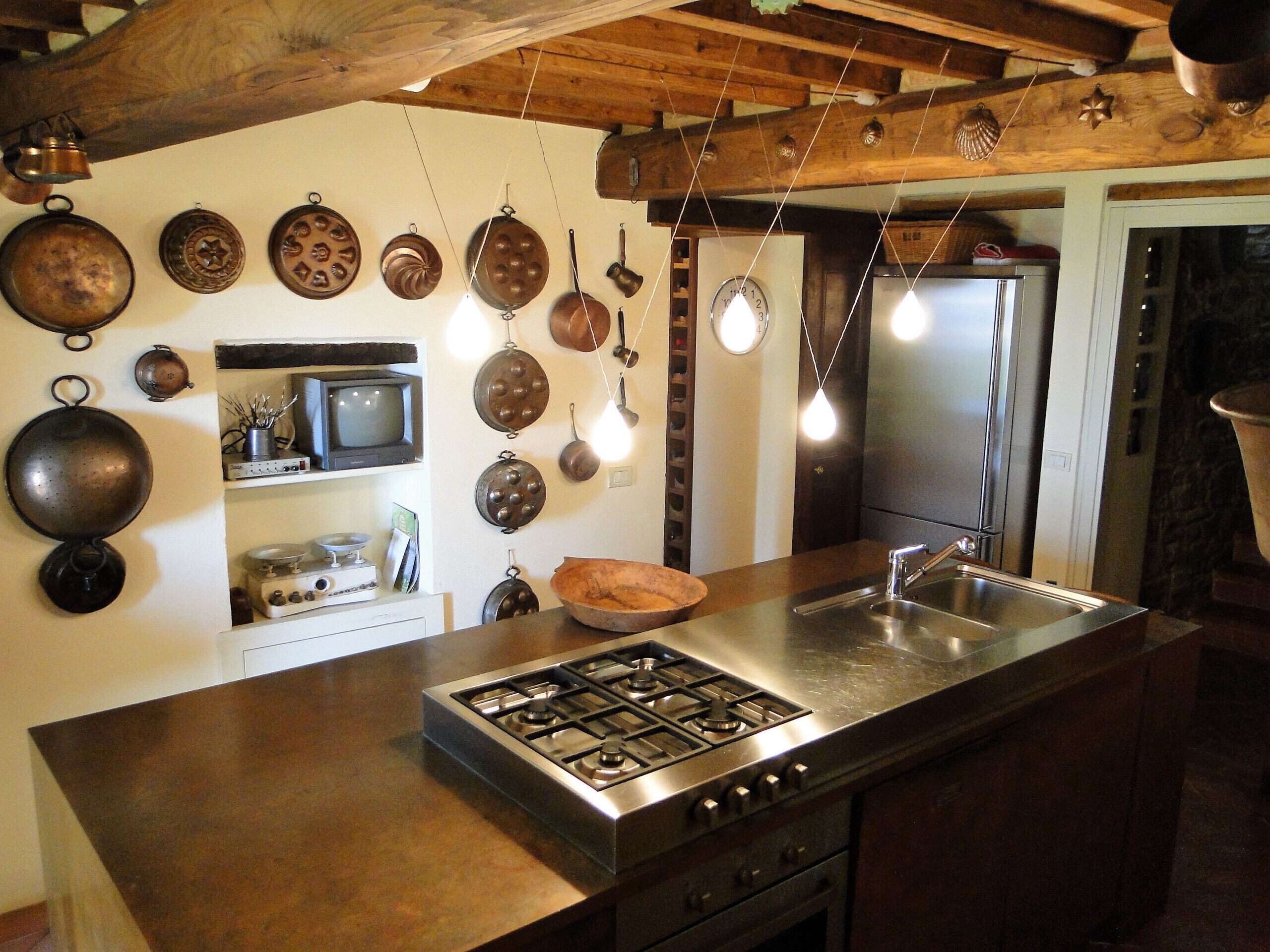 A kitchen with a gas stove, stainless steel sink, wooden ceiling beams, and various hanging pots.