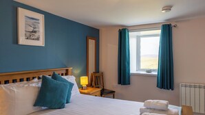 A hotel room with a bed, a nightstand, a chair, a window with curtains, a radiator, and a framed picture on the wall.