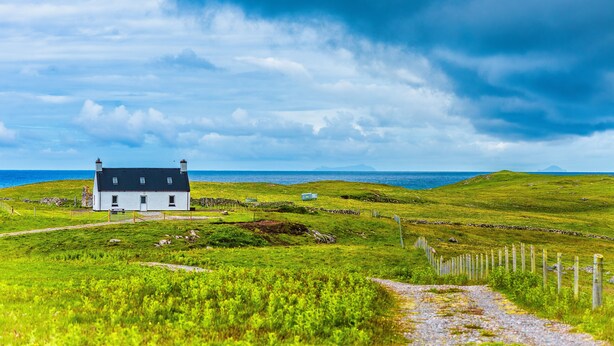 A white house with a dark roof, a stone wall, and a grassy field with a path leading to the sea.