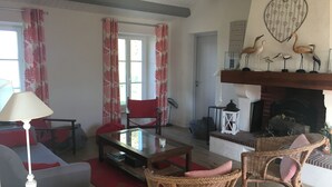 Een woonkamer met een open haard, rieten stoelen, een glazen salontafel en een rood tapijt.