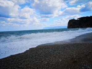 A rocky beach with waves crashing onto the shore, a cliff in the background, and a partly cloudy sky.
