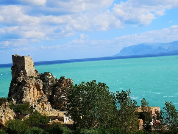 Vista costera con una torre de piedra histórica y edificios, que da a unas aguas turquesas y un cielo despejado.
