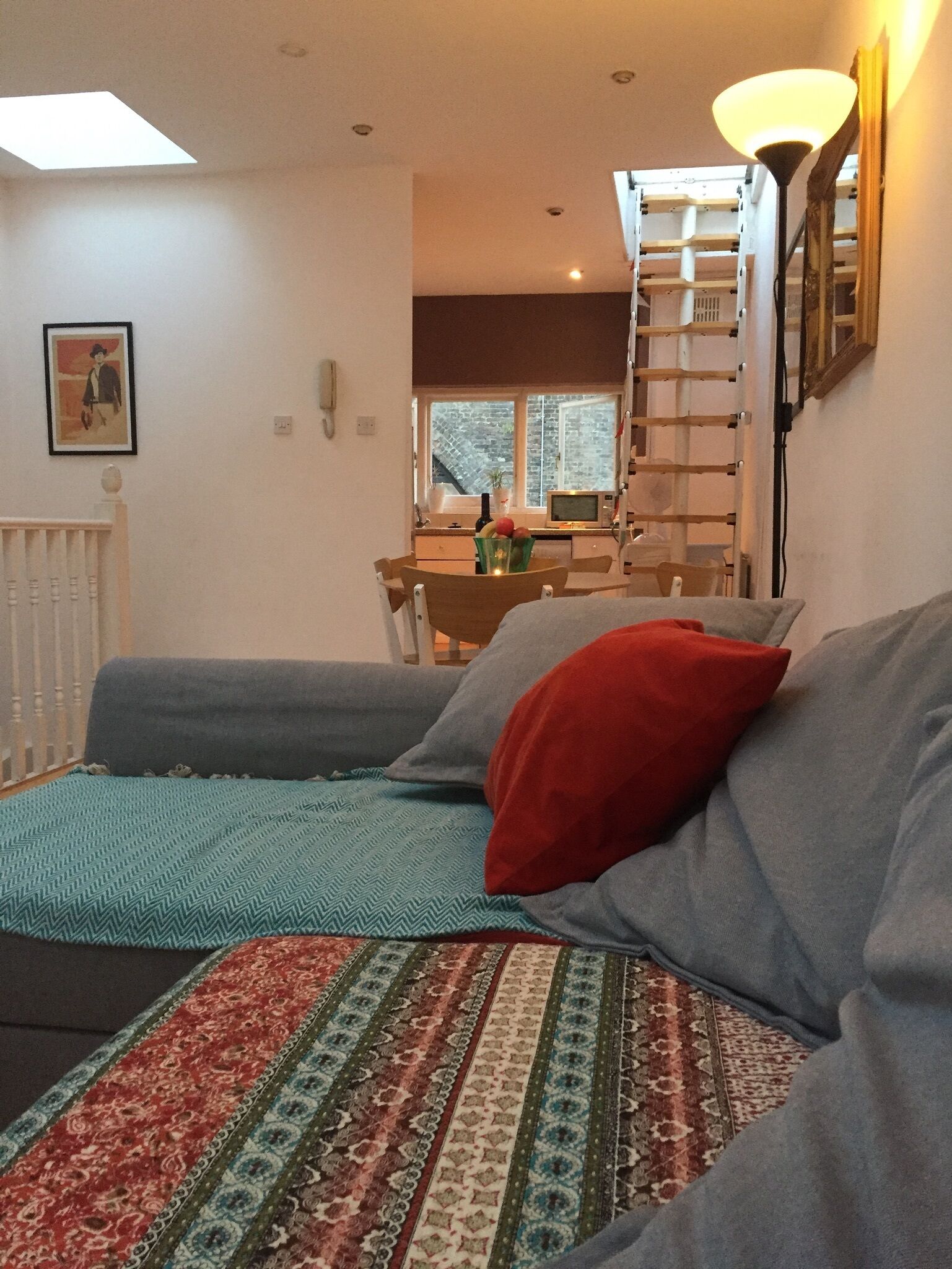 A living room with a sofa, a patterned rug, a red pillow, a ladder, and a window with a view of the outside.