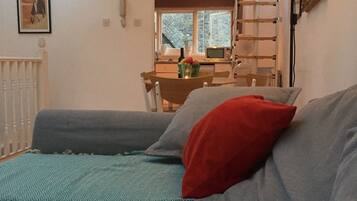 A living room with a sofa, a patterned rug, a red pillow, a ladder, and a window with a view of the outside.