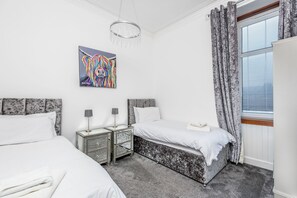 A bedroom with two beds, a nightstand with a lamp, a painting on the wall, and a window with curtains. - Claymore Apartment, Brook Street, central Broughty Ferry, Dundee (Dundee)