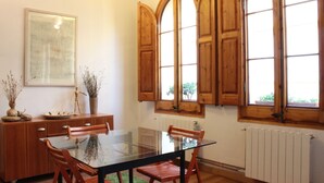 A dining area with a glass table, wooden chairs, a wooden cabinet, and a large window with wooden shutters.