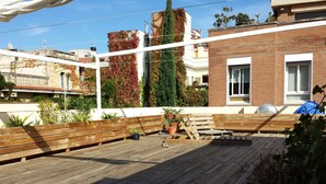 A wooden deck with benches and potted plants, surrounded by buildings with brick and white facades.