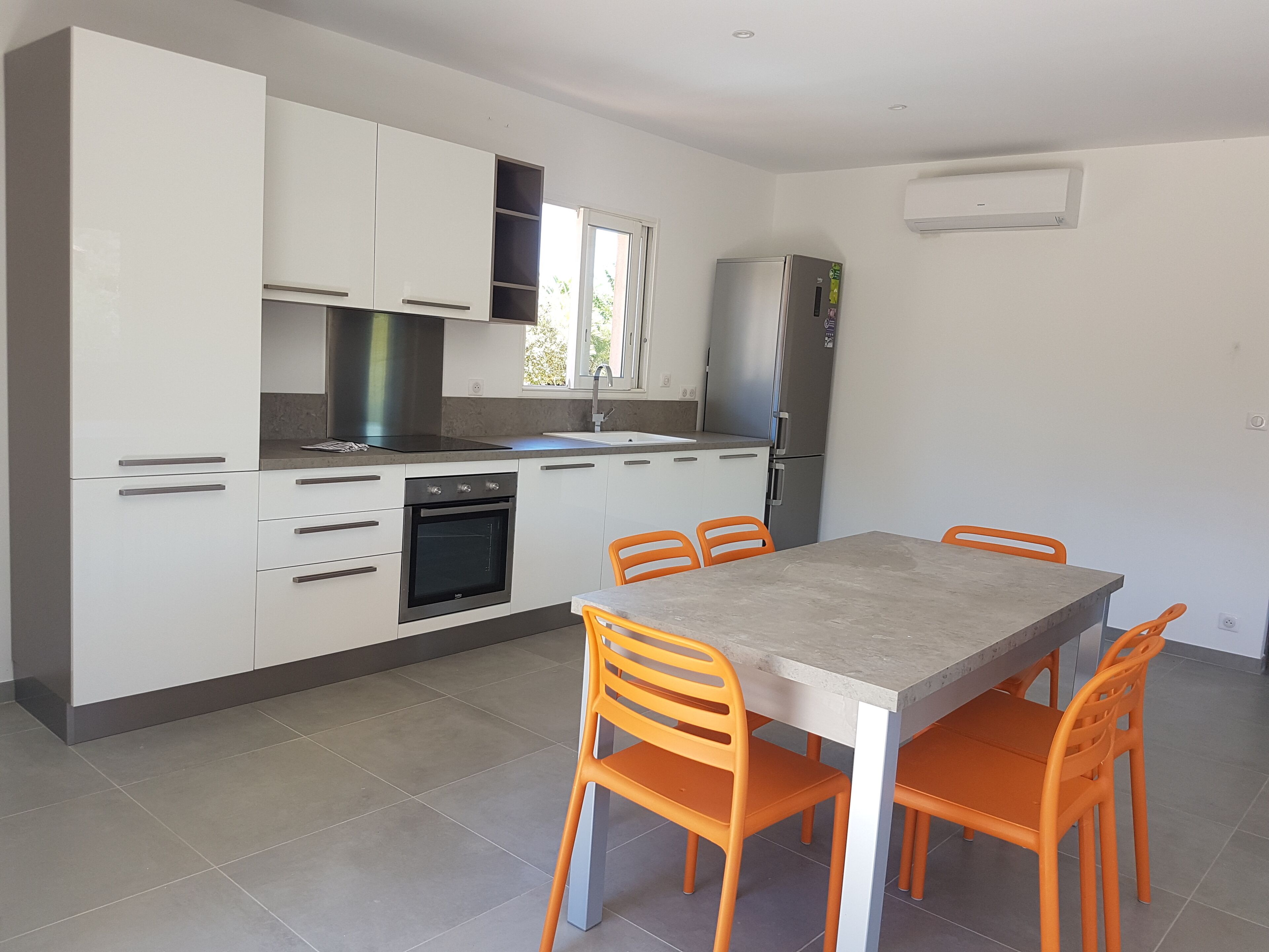 A modern kitchen with white cabinets, a built-in oven, a refrigerator, and a dining table with orange chairs.