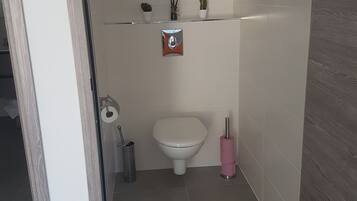 Un baño con inodoro, secamanos, cubo de basura y un estante con objetos decorativos.