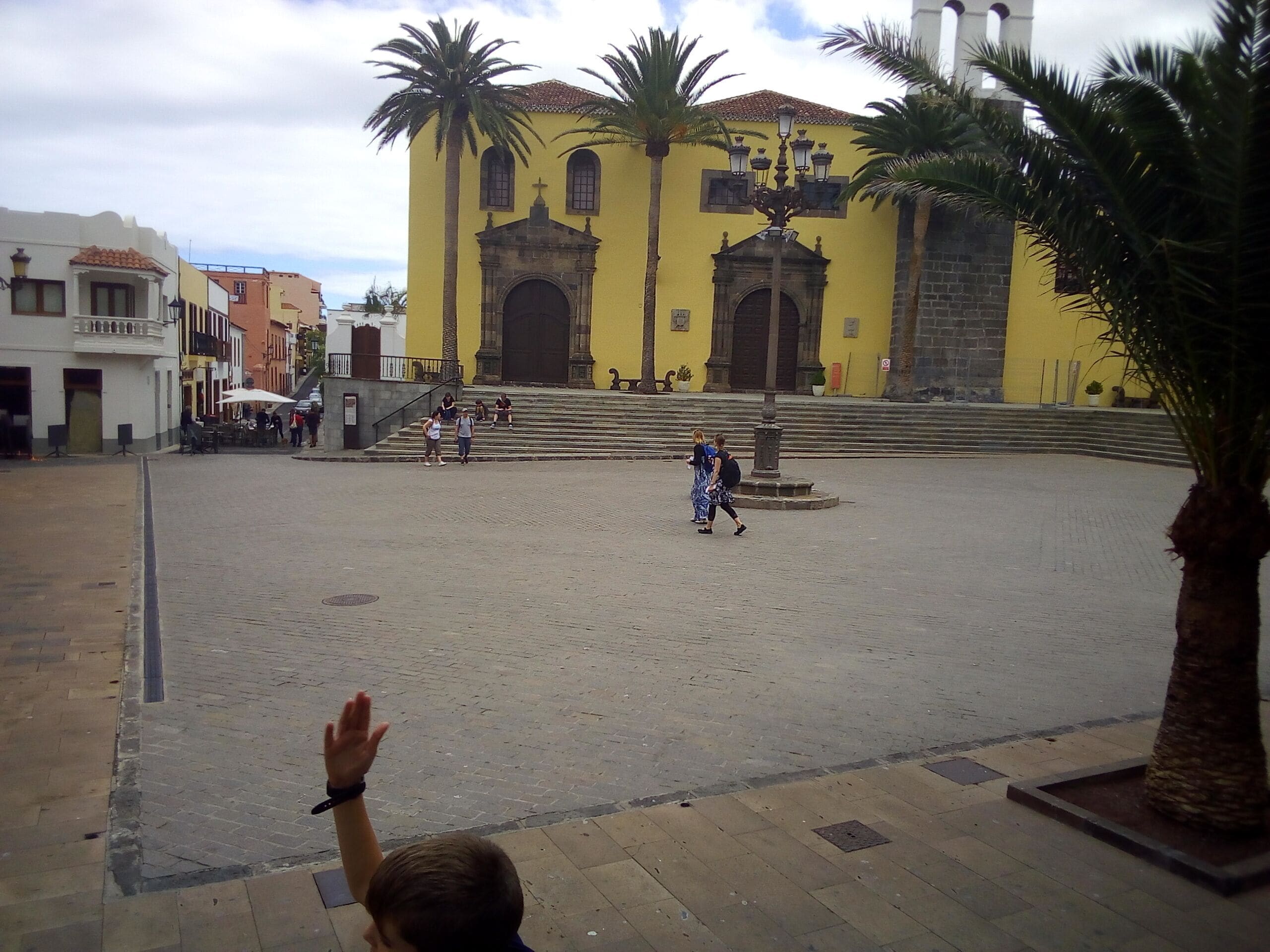 A plaza with a yellow building, palm trees, and a fountain.