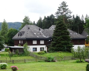 Ein zweigeschossiges Haus mit geneigtem Dach, umgeben von einem Holzzaun und einem Garten mit verschiedenen Pflanzen und Blumen.