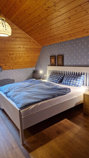 Ein Schlafzimmer mit Holzboden, einem Bett mit kariertem Bettzeug, zwei gerahmten Bildern an der Wand und einem Nachttisch mit Lampe.