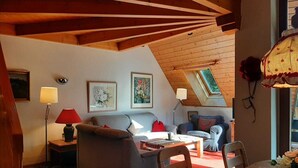 Ein gemütliches Schlafzimmer im Dachgeschoss mit schrägen Wänden, Holzbalken, einem Sofa, einem Esstisch und zwei Lampen.