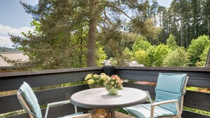 Eine Holzterrasse mit einem runden Tisch und zwei Stühlen, einer Vase mit Blumen und Blick auf Bäume und ein Haus im Hintergrund.