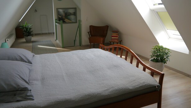 A bedroom with a sloped ceiling, a bed, a chair, a small table with books, and a potted plant. - Art and the sea - close to everything (Varel)