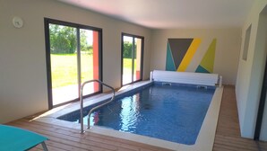 Una piscina interior con cubierta de madera, una escalera metálica y vistas a la vegetación exterior.