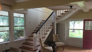 A spacious living area with a wooden staircase, a chair, a small table, and large windows offering views of greenery.