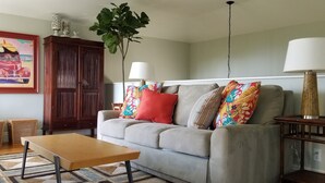Ein Wohnzimmer mit grauem Sofa, einem Holztisch, einem gemusterten Teppich, einer Holzkommode und einem Bild an der Wand.
