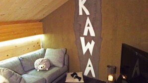 Una habitació acollidora amb un sofà gris, una paret blanca amb les lletres "KAWA" pintades verticalment, una taula petita amb una espelma i un sostre de fusta.