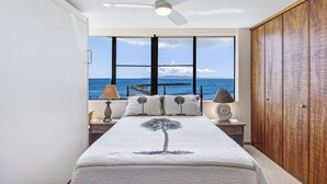 Una camera da letto con un letto, una ventola a soffitto, un comodino con lampade, un'armadio in legno e vista sul mare attraverso ampie finestre.