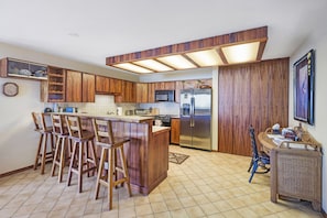 A kitchen with wooden cabinets, a stainless steel refrigerator, a microwave, and a bar area with stools.