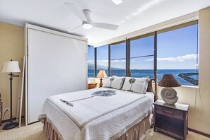 A bedroom with a bed, a ceiling fan, a nightstand, a lamp, and a view of the ocean.