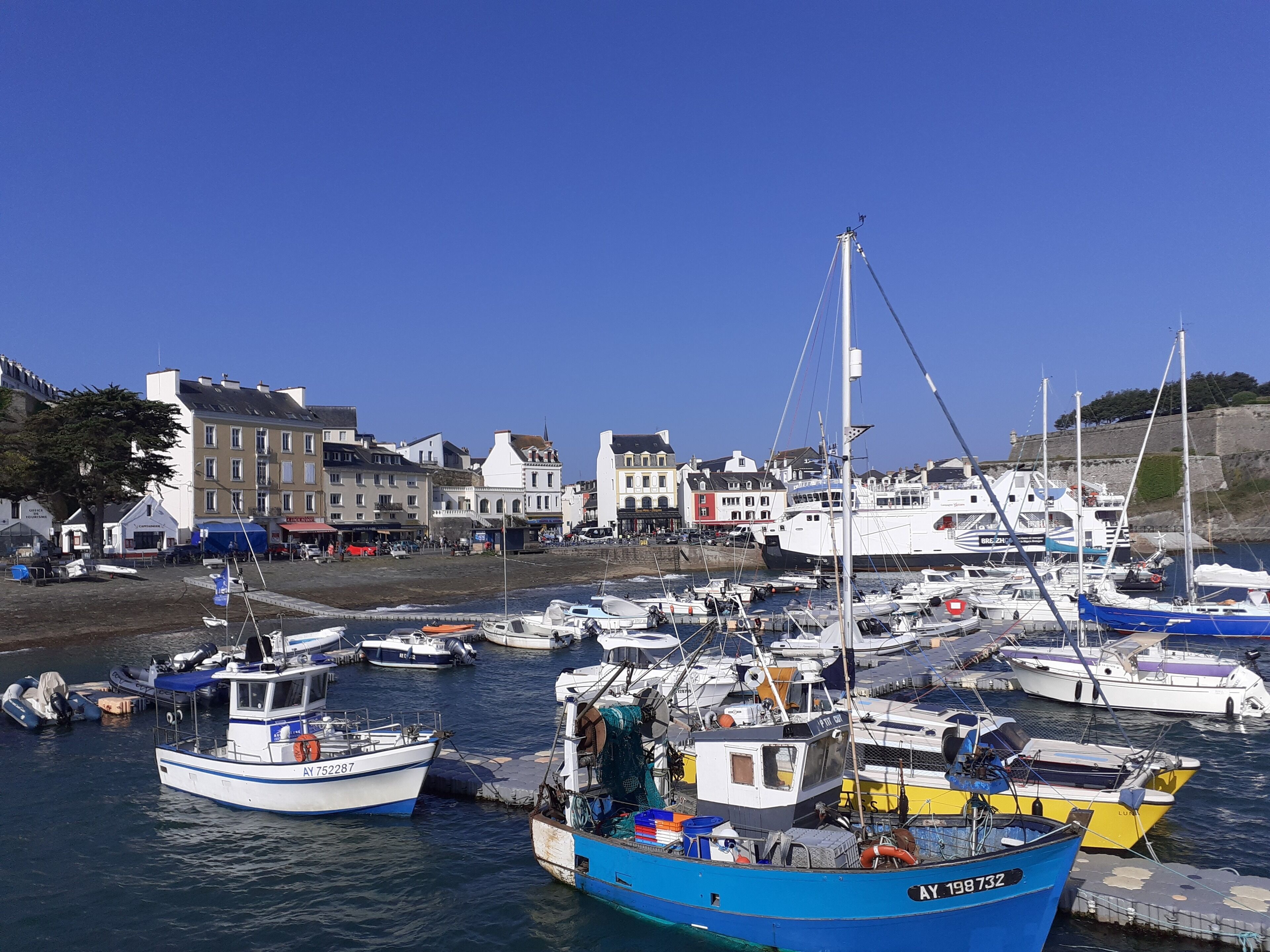 Ein Hafen mit verschiedenen vertäuten Booten, Gebäuden im Hintergrund und blauem, wolkenlosem Himmel.