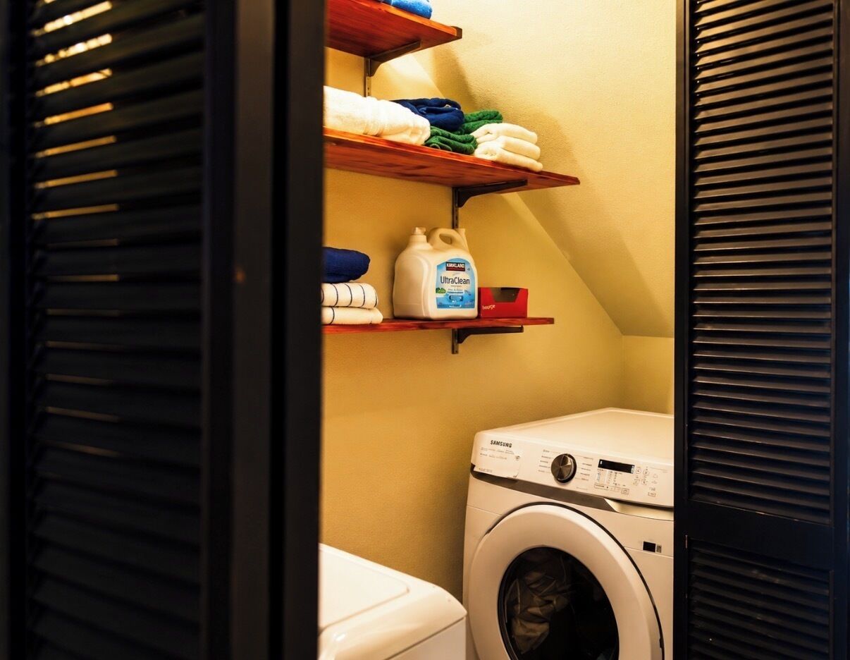 A laundry room with a washing machine, shelves with folded towels, and a bottle of laundry detergent.