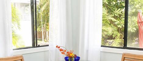 A room with a glass table surrounded by two wooden chairs with dark cushions, a vase with flowers, and a view of greenery through large windows with white curtains.