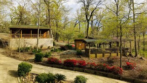 A rustic cabin with a wooden deck and a small porch, surrounded by a landscaped garden with red flowers and various shrubs. - Once a Part of the Beloved Shady Oak Lodge - Cabin 1 - 4 beds with Boat Ramp (Many)