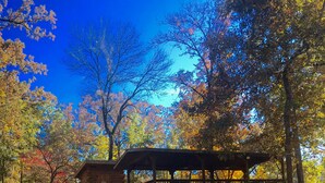 A wooden gazebo with a covered seating area, surrounded by trees with autumn foliage, and a small building with a distinctive sun-shaped window. - Once a Part of the Beloved Shady Oak Lodge - Cabin 1 - 4 beds with Boat Ramp (Many)