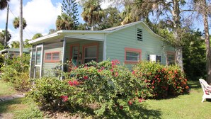A light green house with a porch, surrounded by lush greenery and flowering bushes.