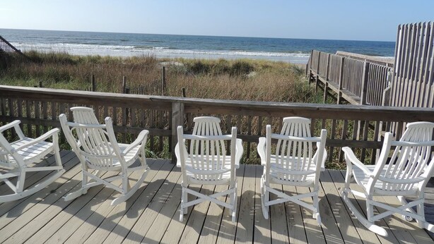 Een houten terras met witte Adirondack-stoelen gericht op het strand.