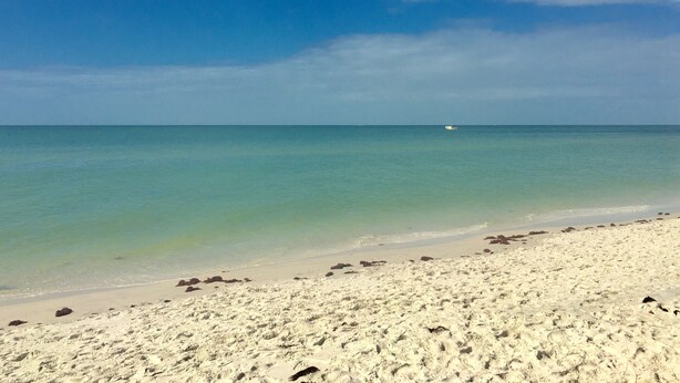 En strand med klart turkis vand, en båd i det fjerne og en sandet kystlinje.