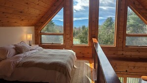 A wooden cabin bedroom with a bed, a nightstand, and a lamp. There are large windows offering a view of the forest and mountains. - 5BR Catskills Retreat • Ski Close • Hot Tub, Firepit, 2 grills, Deck, Fast WiFi (Prattsville)