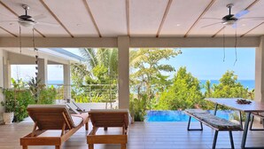 A spacious outdoor area with wooden lounge chairs and a pool table, overlooking a clear blue sea and lush greenery.