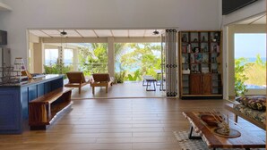 A spacious living area with a wooden floor, a kitchen counter, and a seating area with a sofa and chairs. There are two windows providing natural light and a view of the outdoors.